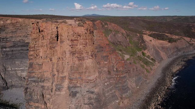 Man standing on stratified sandstone cliff at Vila Nova de Mil Fontes Alentejo Portugal above Atlantic Ocean highlighting scale of dramatic fal&eacute;sia and coastal erosion landscape, drone orbiting
