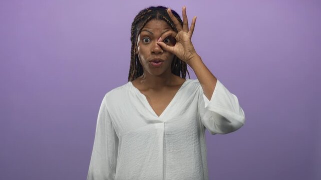 Woman wearing white blouse making ok circle hand pose over eye in soft purple studio; playful curiosity.
