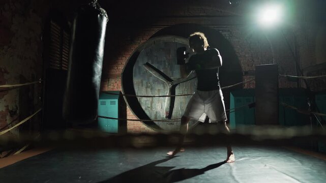 Fighter practicing hands punches in boxing ring, motivated sportsman, portrait. Full-length shot with professional athlete training in gym in night, preparing to important competition, hard workout
