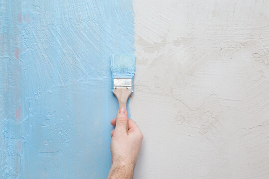 Young adult man hand holding brush and applying blue waterproofing coating on concrete wall or floor before lying tiles at bathroom. Closeup. Repair work of home. Front view.