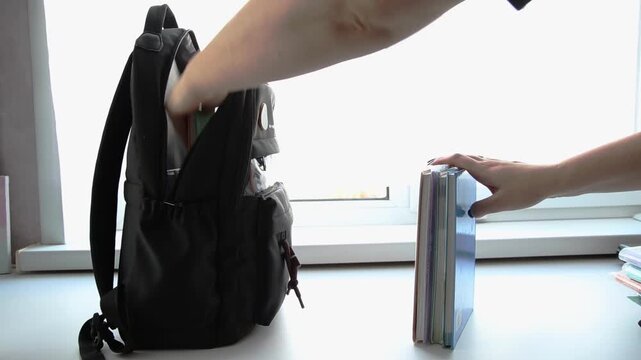 Person hands packing black backpack and organizing stack of books on white surface next to bright window. Scene depicts student preparation for school or study.