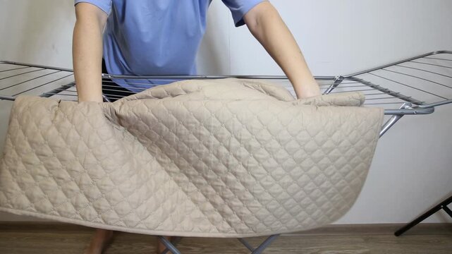 Person carefully positioning clean quilted beige mattress cover on metal drying rack indoors. Focus on domestic chore, routine laundry activity, fresh linen care, textile diamond pattern.