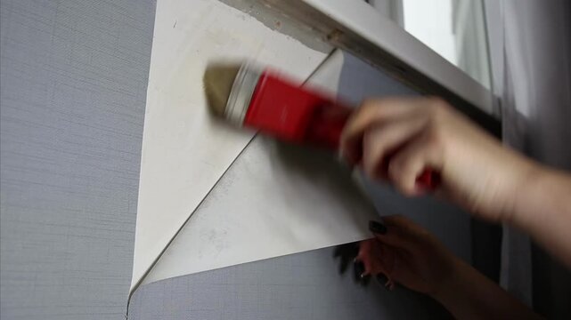 Close up view of person hand using red brush to smooth new wallpaper onto grey wall. Home improvement activity with adhesive application and surface preparation.