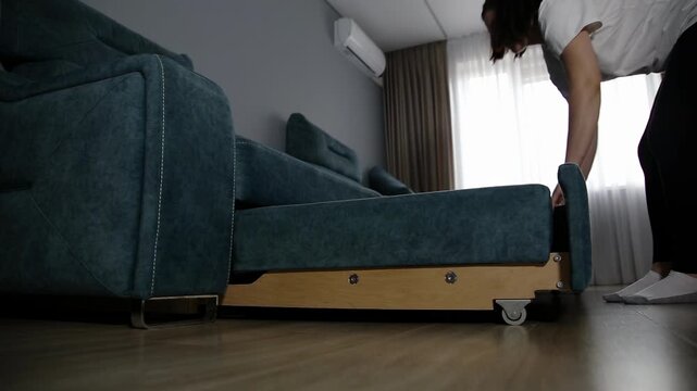 Woman converts teal sleeper sofa to bed. Action demonstrates furniture transformation, revealing mechanism. Modern space saving solution, home interior comfort.