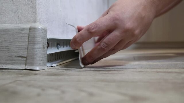 Hands install plastic baseboard cover along interior wall. Cable management system hides wiring. Home renovation on wood effect laminate floor.