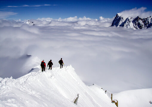 Three climbers ascend a snowy mountain ridge above a sea of clouds, braving the elements for the summit.