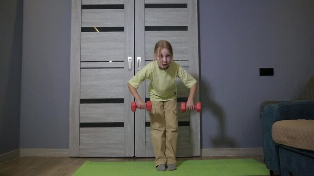 Young blonde girl concentrates on fitness routine, performing bent over rows with red dumbbells on green yoga mat. Girl wears yellow t-shirt and beige pants, exercise indoors.