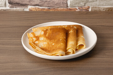 Warm crepes served on a white plate placed on a wooden table with a brick wall background during daytime