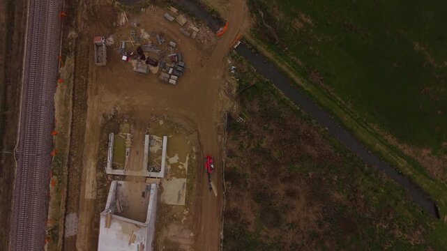 Drone perspective of a railway construction site in Milan showing workers, machinery and earthworks during spring, with cloudy weather creating a muted industrial setting.