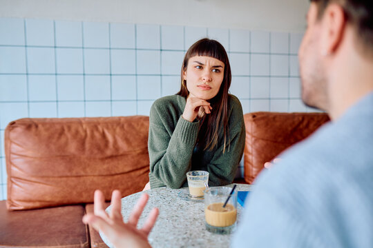 Woman looking with a skeptical facial expression, listening to a man talking, sharing coffee drinks in a modern cafe