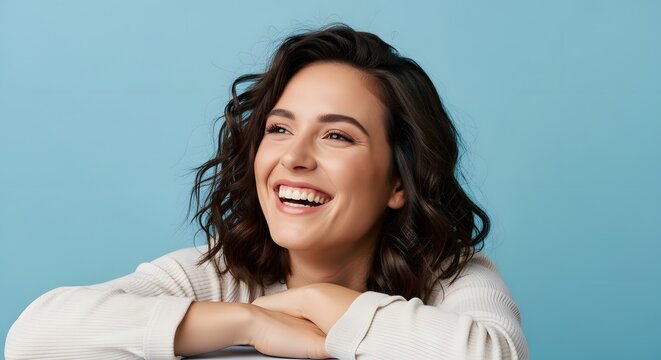 Cheerful young woman with wavy brown hair laughing, happy female portrait on blue background, joyful person expressing positivity and wellness