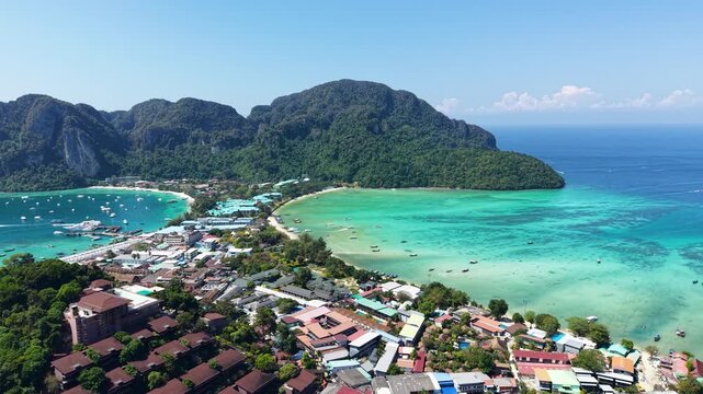 Rotating aerial drone view of Phi Phi Island village between two turquoise bays in Krabi, Thailand. Tropical beaches, boats and lush limestone mountains create a stunning island landscape.