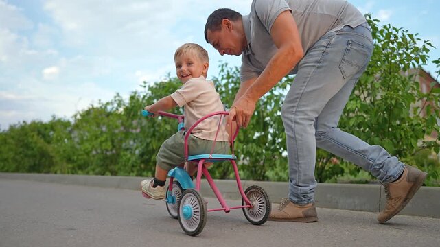 Happy father pushes little son on tricycle along summer sidewalk. Dad helps child ride bike outdoors. Parent teaches boy to steer. Family enjoys active day. Father loves guiding son on tricycle.