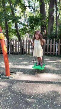 Vertical video of happy little girl having fun on the playground. Swinging on swing. 