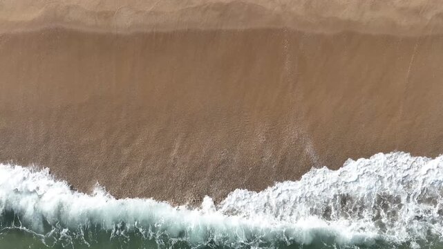 Aerial view of ocean waves crashing onto a sandy beach. Natural texture, summer vibes, and scenic coastal beauty.