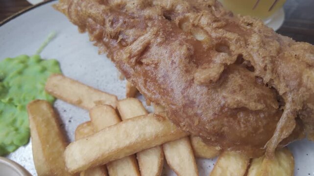 Close up of a classic plate of British fish and chips, featuring crispy battered fish, thick cut french fries, mushy peas, and tartar sauce served on a rustic plate in a restaurant