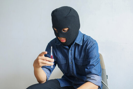 Masked Man Sits Holding Syringe Filled with red Liquid, Representing Drug Addiction