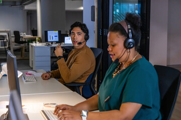 Diverse coworkers working side-by-side at open-plan office desk, wearing headsets, viewing charts © wavebreak3