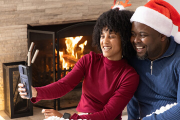 Naklejka premium African American couple taking selfie in living room by lit fireplace with smartphone and Santa hat