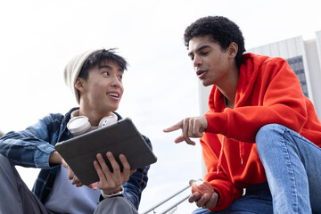 Obraz premium Diverse male teenagers sitting on campus steps, pointing at tablet, holding apple and headphones