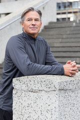 Senior man leaning on concrete post in city plaza near stairway, wearing gray jacket, looking right © wavebreak3