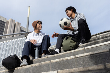 Diverse teen males sitting on concrete steps wearing hoodies holding soccer ball and backpacks © wavebreak3