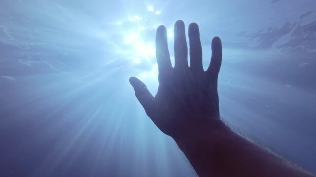 POV underwater video: Man's hand waving in greeting to bright dancing sun rays, backlit by sunlight. Morning god rays pierce surface of water, man's hand waves in greeting to new day in under water