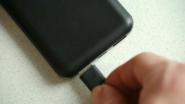 Close up hand plugging black usb cable in port portable power bank, recharging external battery charger to provide power for mobile electronic devices on go. Connecting usb cable to black power bank