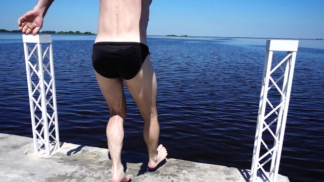 Athletic Young Man in Swim Briefs Standing on the Edge of a Concrete Pier Overlooking a Vast Blue Lake