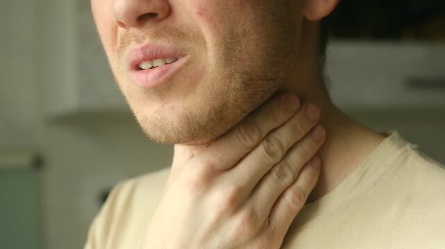 Close up man clutching his neck in pain, suffering from sore throat, tonsillitis, or other respiratory illness, grimacing from discomfort of inflammation and difficulty swallowing. 