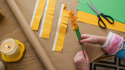 Child's hands crafting a paper flower with yellow and green materials on a table