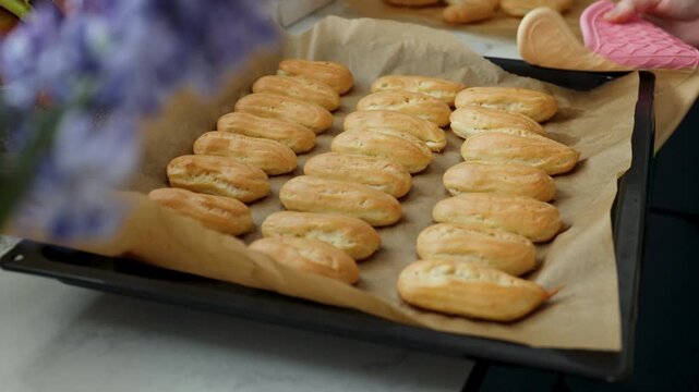 Baker handling golden choux pastries on baking sheet. Fresh baked shells for custard eclairs in sunny kitchen. Home dessert preparation process with oven mitt. video footage video footage photography