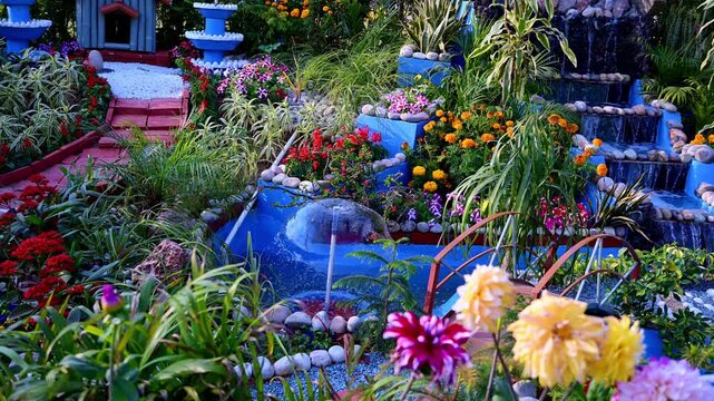 Beautiful landscaped garden with colorful flowers, cascading waterfall fountain, decorative bridge, and small water feature surrounded by lush plants and stones.