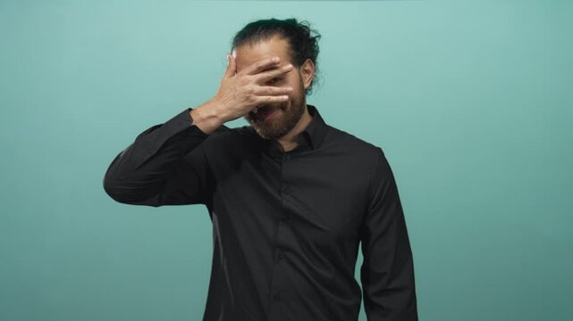 Young man with beard wearing black shirt covering eyes with hand in teal green studio; shyness curiosity.