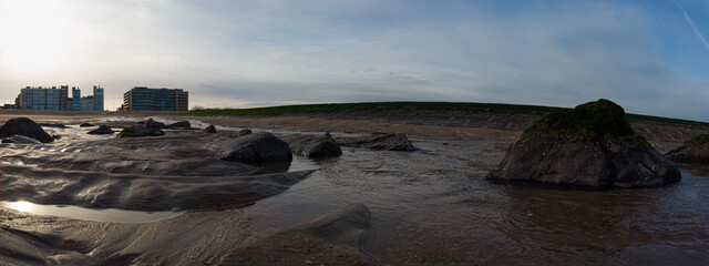 Low tide exposes wet sand, rocks and shallow tide pools along a coastal shoreline with a grassy sea...