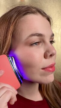 A young woman smooths her facial skin with a special electric massager during her morning hygiene routine. Skin care concept