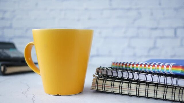 Yellow ceramic cup on desk next to stack of spiral notebooks in office setting with cinematic slow camera movement