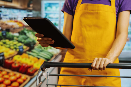Grocery store worker in yellow apron holds tablet over shopping cart in produce aisle, checking inventory or online order fulfillment, with fresh vegetables in background.