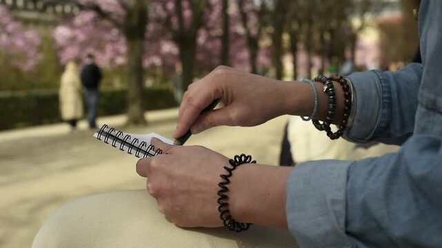 Close up of a woman's hands sketching pink magnolia flowers in a spiral notebook at the Luxembourg Garden in Paris during spring.