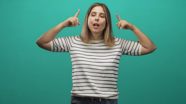 Woman covering ears with hands and rolling eyes, wearing striped white shirt and jeans in studio teal backdrop; annoyance blocking noise.