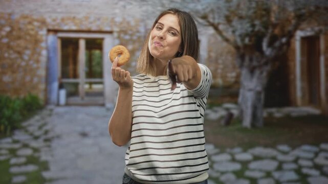 Woman holding a donut and pointing finger at camera in a stone courtyard beside a building, wearing striped shirt and smiling with crumbs on lips; playful confidence.