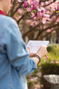 Close up of a woman's hands sketching pink magnolia flowers in a spiral notebook at the Luxembourg Garden in Paris during spring.