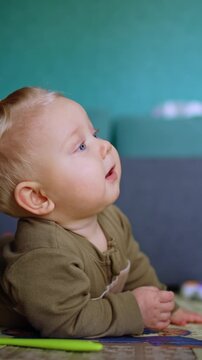 Baby with lovely plump cheeks lies on the floor looking up with attention. Infant moves by the floor crawling. Vertical video