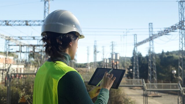 Female engineer reviewing data on a digital tablet at an electrical power substation, ensuring infrastructure maintenance and managing the transmission of energy and utilities