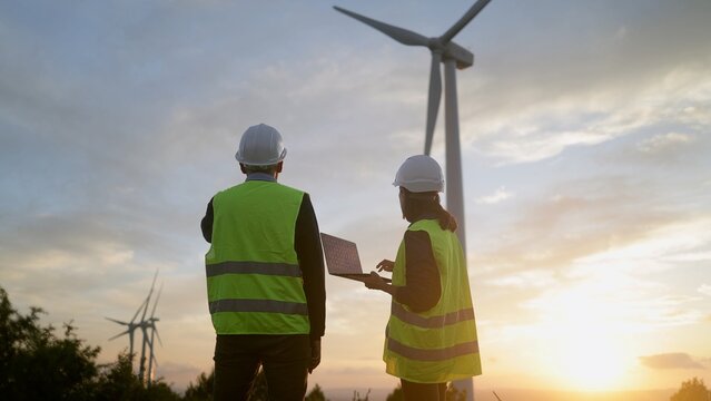Engineers with hard hats and high visibility vests inspecting a wind farm at sunset, working on clean energy generation and discussing sustainability development