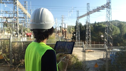 Engineer holding a tablet displaying data and using a stylus, overseeing operations at an active power station with high voltage electricity pylons and transmission lines