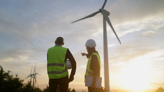 Engineers in safety vests overlooking a wind turbine farm at sunset, discussing renewable energy development and sustainability for a greener future