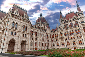 Obraz premium The Hungarian Parliament Building stands under a dramatic, colorful sky with intricate Neo-Gothic architectural details and a central dome.