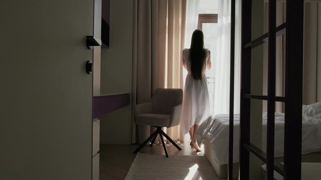 Woman closing curtain and walking away from window in minimalist bedroom interior. Modern apartment with loft bed above sofa, soft natural light and neutral color palette. 