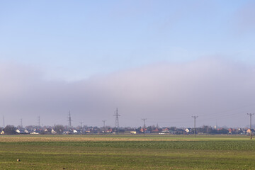Fototapeta premium Village houses and prominent electricity pylons emerge from low-lying mist across green fields, highlighting rural energy infrastructure under a clear sky